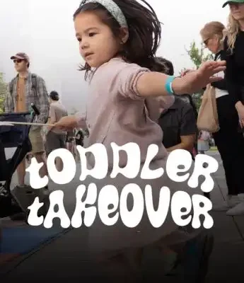 Young girl in mid-motion with arms outstretched, surrounded by adults outdoors. Text overlay reads "Toddler Takeover" as she joyfully leads the group.
