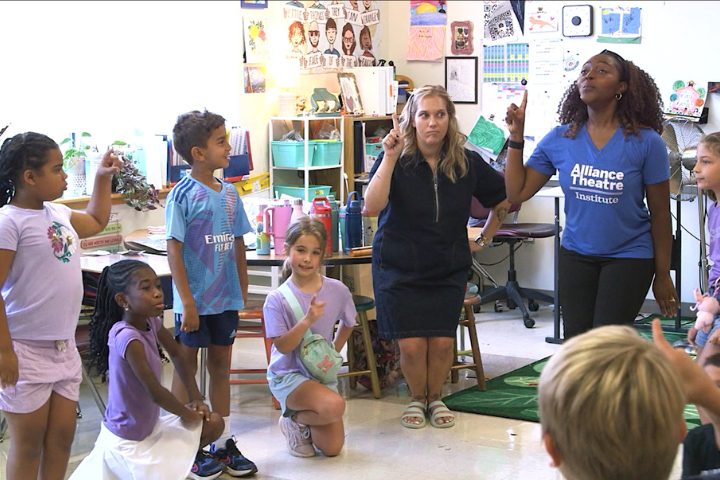 A group of children and two adults participate in an activity in a colorful classroom, some standing and some kneeling, with their fingers raised as if signaling or gesturing.