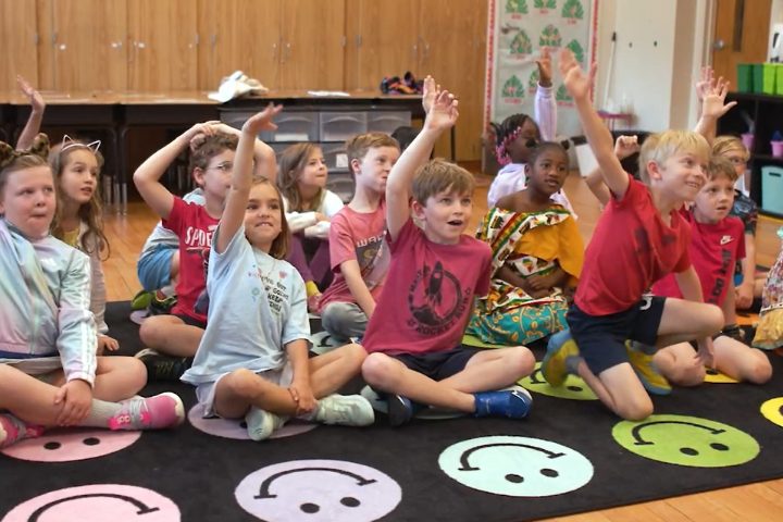 A group of young children sit on a colorful rug in a classroom, raising their hands and looking toward the front of the room.