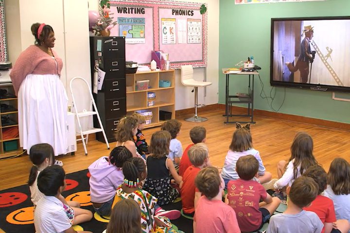 A teacher stands at the front of a classroom while a group of young students watch a video featuring a person using a ladder on a television.