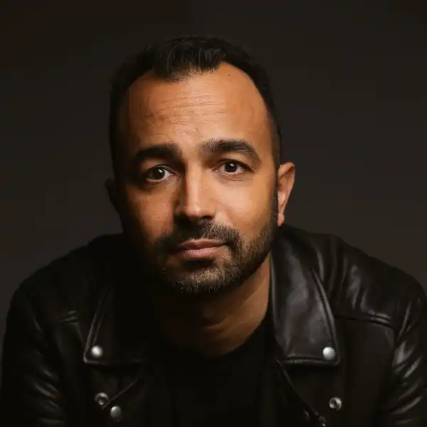 Man with short dark hair and beard wearing a black leather jacket, looking directly at the camera against a dark background.