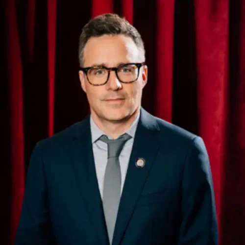 A man wearing glasses and a blue suit with a tie stands in front of dark red curtains.