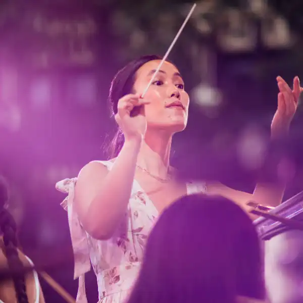 Cynthia Meng, in a light floral dress, conducts an orchestra with a raised baton under purple stage lighting.