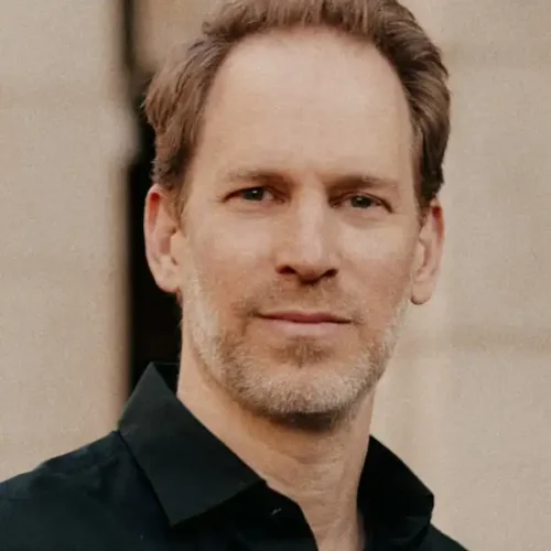 A man with short light brown hair and light facial stubble, wearing a black collared shirt, stands in front of a neutral background.