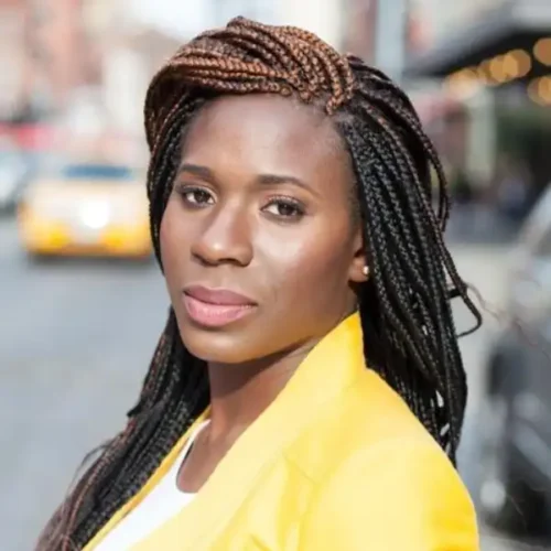 A woman with long, braided hair wears a yellow blazer and looks at the camera while standing on a city street.