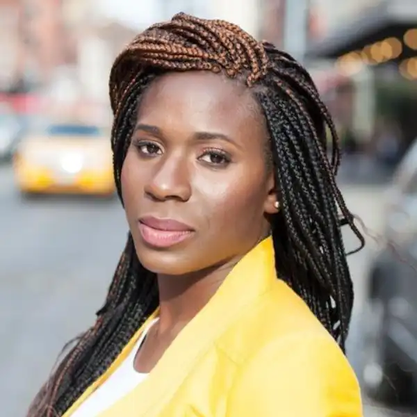 A woman with long, braided hair wears a yellow blazer and looks at the camera while standing on a city street.