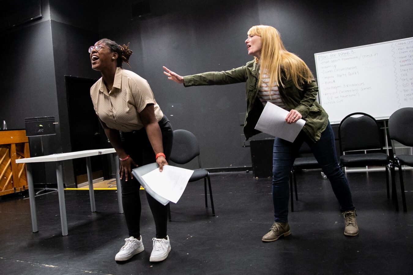 Two women rehearse a scene on stage, holding scripts; one leans forward, laughing, while the other reaches out toward her. A whiteboard and chairs are visible in the background.