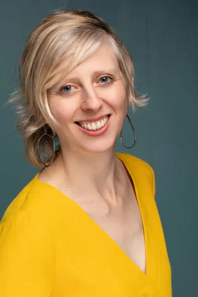 Smiling person with short blonde hair wearing a yellow top and large hoop earrings, posed against a plain teal background.