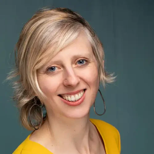 Smiling person with short blonde hair wearing a yellow top and large hoop earrings, posed against a plain teal background.