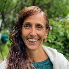 Victoria Navarro, a woman with long brown hair, wearing a green sweater and a light-colored shawl, smiles outdoors with lush greenery in the background.