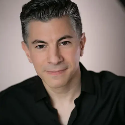 A man with short, dark hair and wearing a black shirt looks directly at the camera with a neutral expression against a light background.