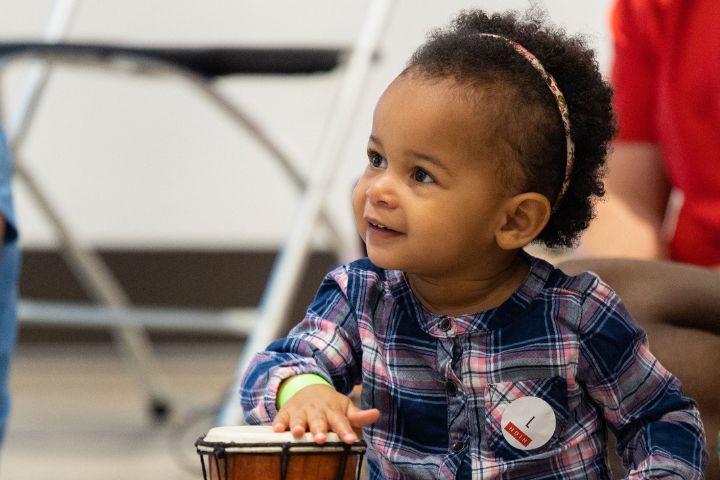 A young child in a plaid shirt sits indoors, smiling and tapping on a small drum.