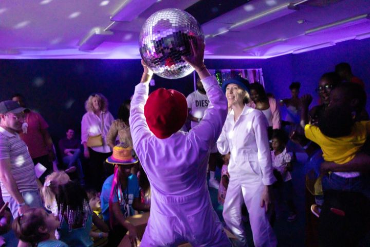 A person in a red hat holds up a disco ball in a room filled with people, some in costumes, under purple lighting with reflected light spots.