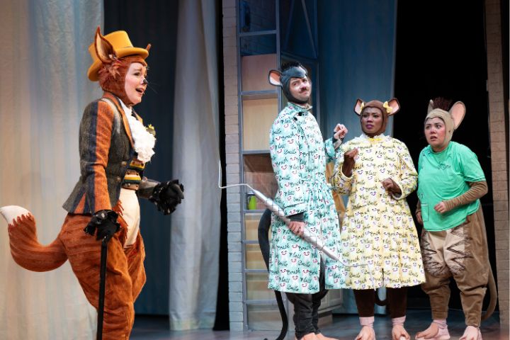Four actors in animal costumes, including a fox and three mice, perform on stage; the set includes a metal ladder and a white curtain in the background.