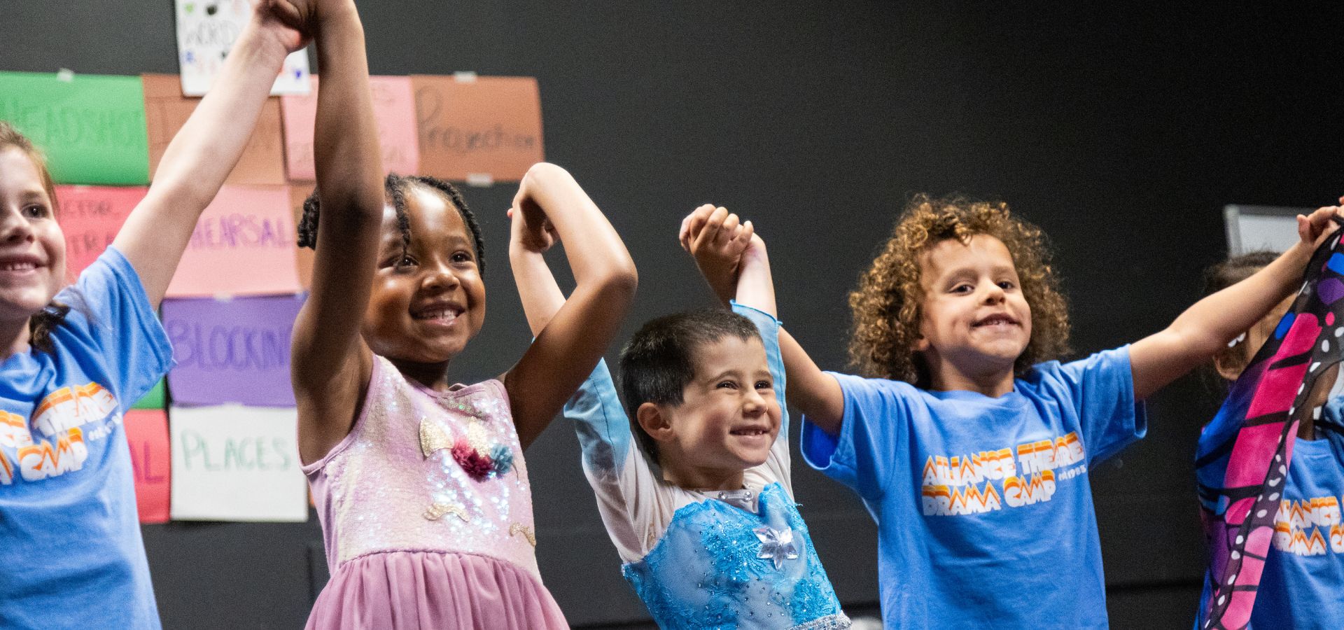Four young children, smiling and holding hands in the air, stand together in costume and blue t-shirts in front of a wall with colorful paper signs at Alliance Theatre drama camp.