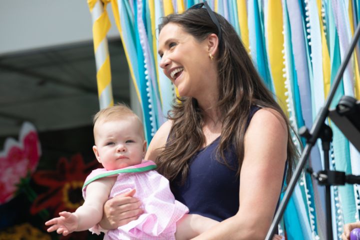 A woman holding a baby stands in front of colorful ribbons, smiling, with a microphone visible in the foreground.