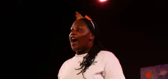 High school student in a white shirt and orange headband stands under stage lights at musical theater camp, appearing to sing with an open mouth against a dark background.