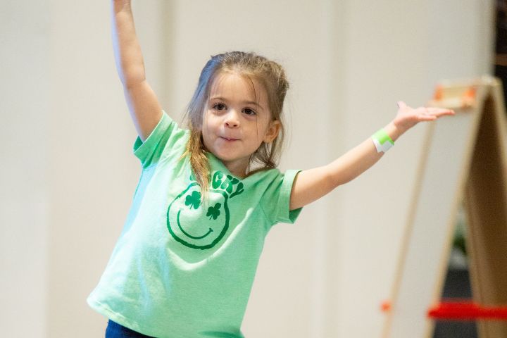 Young girl wearing a green T-shirt with a smiling face graphic stands with arms raised, smiling indoors.