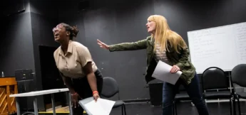 Two women hold scripts while rehearsing in a theater rehearsal room in Viewpoints class; one leans forward, laughing, as the other extends a hand toward her.