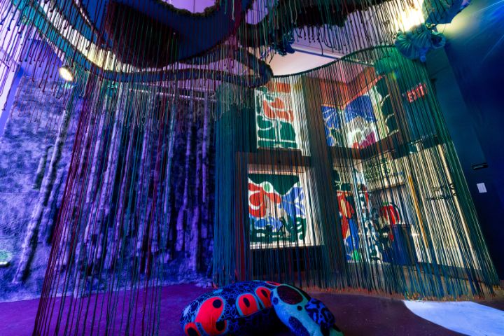 Room with hanging strands of colorful string, abstract patterned walls, and a soft, polka-dotted sculptural seat on the floor, illuminated by blue and purple lighting.