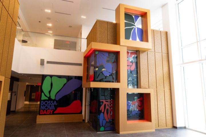 Modern building interior with tall windows, artwork featuring bold abstract shapes and colors, and tan wall panels. The text "Bossa Nova Baby" appears on the lower left artwork.
