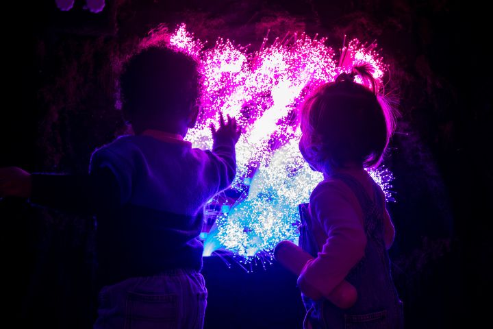Two young children stand in front of a glowing, colorful light display with pink, purple, and blue hues in a dark setting.