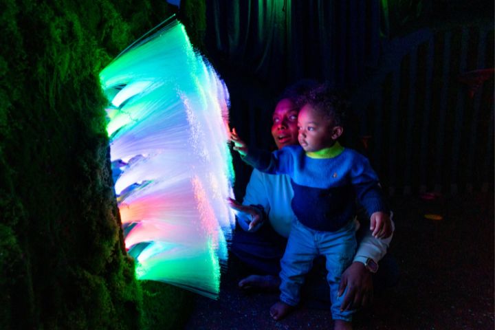 An adult and child touch glowing fiber optic lights in a dark, indoor interactive exhibit.