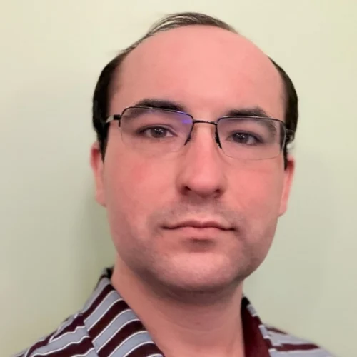 A man with short dark hair and glasses is wearing a maroon and blue striped collared shirt, facing the camera against a plain light-colored background.