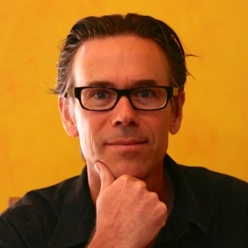 Colin Gagné, wearing black glasses and a black shirt, sits in front of a yellow background, resting his chin on his hand and looking at the camera.