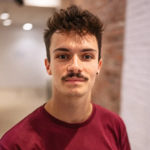 A young man with short brown hair and a mustache, wearing a maroon shirt, stands indoors near a brick wall, looking at the camera.