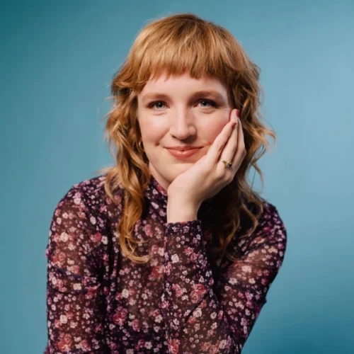 A person with wavy, shoulder-length reddish-blonde hair sits against a blue background, wearing a dark floral patterned top and resting their face on their hand, smiling faintly.