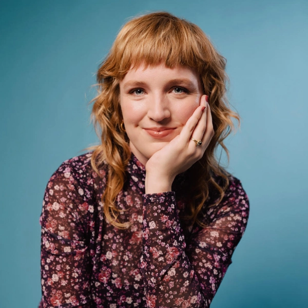 A person with wavy, shoulder-length reddish-blonde hair sits against a blue background, wearing a dark floral patterned top and resting their face on their hand, smiling faintly.