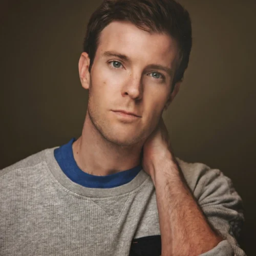 A young man with short brown hair wears a grey sweatshirt and blue shirt, looking at the camera with his hand resting behind his neck against a neutral background.