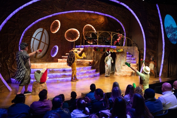 Actors in colorful costumes perform a stage play with a fantasy set design, while an audience—some winners of the digital lottery—watches from their seats.