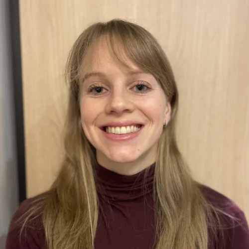 A person with long blonde hair wearing a maroon turtleneck smiles at the camera, standing in front of a light wooden background.