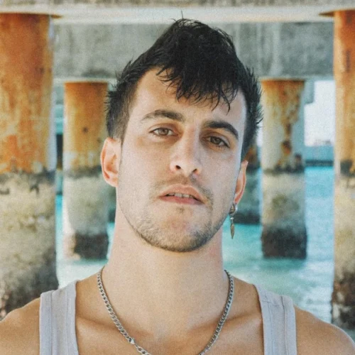 A man with short dark hair, a light beard, and a silver chain necklace stands under a pier by the water, wearing a gray tank top.