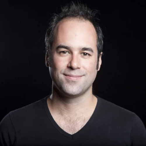 Colin Gagné, a man with short dark hair and a slight smile, is wearing a black V-neck shirt against a plain black background.