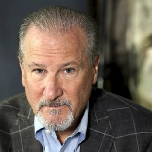 An older man with gray hair and a goatee, wearing a gray plaid suit jacket and light blue shirt, looks directly at the camera.