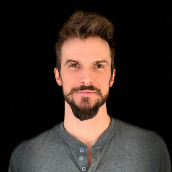 A man with short brown hair, a styled beard, and a mustache is wearing a gray henley shirt and facing the camera against a plain black background.