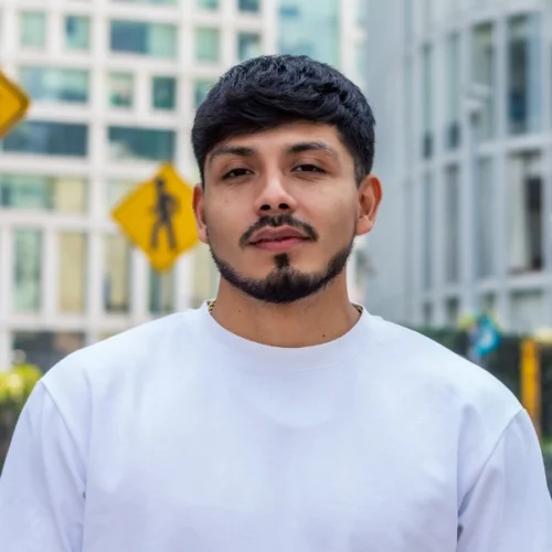 A man with short dark hair and a trimmed beard wears a white shirt and stands outdoors in an urban setting with blurred buildings and road signs in the background.