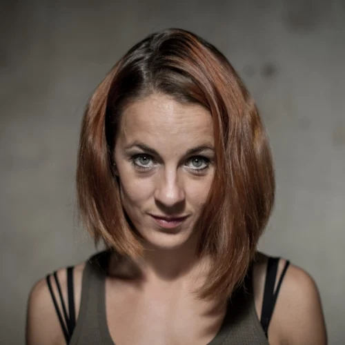 Héloïse Bourgeois, with shoulder-length auburn hair and green eyes, looks directly at the camera, wearing a sleeveless top with thin straps against a neutral background.