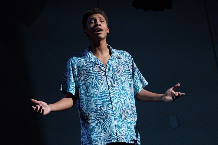 Isaiah C. Phipps wearing a patterned short-sleeve shirt stands under stage lighting with arms outstretched, appearing to perform or sing performing in the Palefsky Collision Project.