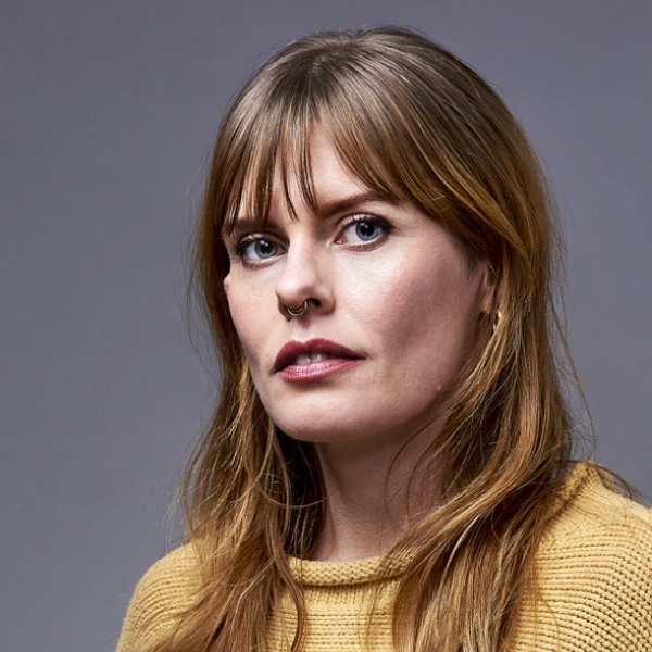 A woman with long light brown hair, wearing a yellow sweater and a nose ring, looks directly at the camera in front of a plain gray background.