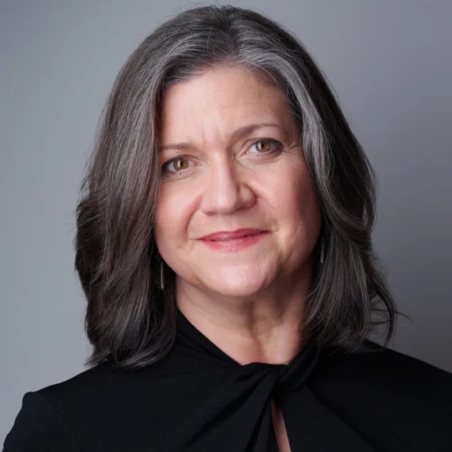A woman with shoulder-length gray hair and a black top is smiling slightly in front of a plain gray background.
