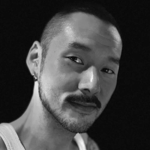 A person with short hair, a mustache, and a beard stubble looks at the camera in a black and white portrait, wearing a tank top and an earring.