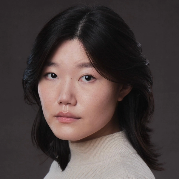 A woman with medium-length dark hair wearing a light beige turtleneck top, looking at the camera with a neutral expression against a dark background.