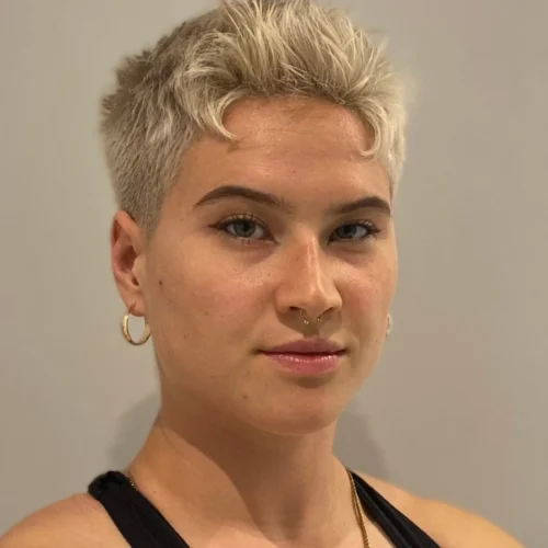 A person with short, platinum blonde hair, wearing hoop earrings and a nose ring, looks directly at the camera against a plain background.