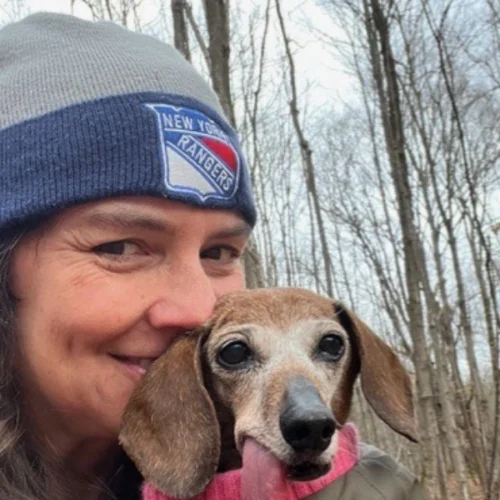 Madeleine Oldham smiles wearing a New York Rangers beanie next to a brown dachshund dog with its tongue sticking out, outdoors with leafless trees in the background.
