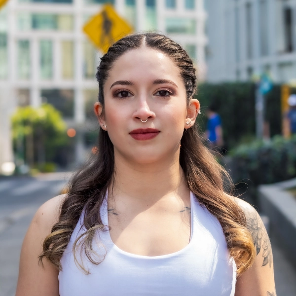 A woman with long brown hair and a nose ring stands outdoors in a city, wearing a white sleeveless top.