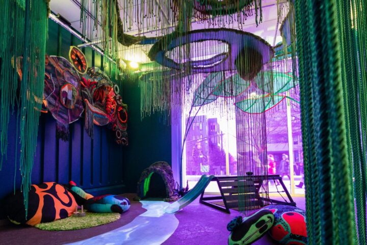 Colorful, immersive PNC PlaySpace with hanging green strands, large fabric art pieces, beanbags, a small slide, and purple lighting near a window.